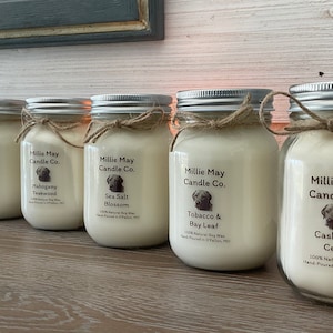 May include: Six glass jars with silver lids and twine bows. Each jar contains a soy wax candle with a different scent. The labels on the jars read "Millie May Candle Co." and list the scent of each candle: "Raspberry Vanilla", "Mahogany Teakwood", "Sea Salt Blossom", "Tobacco & Bay Leaf", and "Cashmere Cedar".