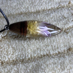 May include: A brown wooden pendant with a clear resin insert containing a pressed purple flower. The pendant is attached to a black cord.