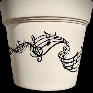 May include: Cream-coloured ceramic plant pot featuring a black musical note design. The pot has a gently flared shape with a decorative band near the top. The musical notes are stylised and flow across the pot's surface.