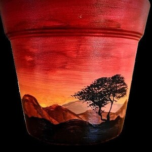 May include: A terracotta flower pot painted with a sunset scene. The pot features a silhouette of a tree and mountains against a red, orange, and yellow sky.