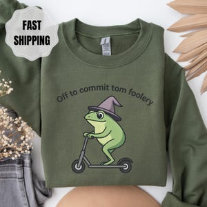 Könnte beinhalten: Olivgrünes Sweatshirt mit einer Cartoon-Fröschin, die einen Hexenhut trägt und einen Roller fährt. Der Text "Off to commit tom foolery" ist über dem Frosch. Das Sweatshirt ist aus einem weichen Material.