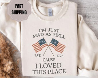 May include: Sand-colored sweatshirt with the text "I'M JUST MAD AS HELL CAUSE I LOVED THIS PLACE" and two crossed American flags. The sweatshirt also has the text "EST. 1776".
