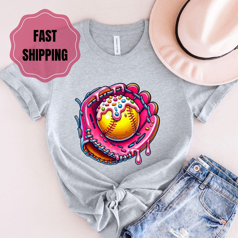 May include: A gray t-shirt with a pink and yellow graphic of a baseball in a catcher's mitt with pink frosting and sprinkles. The graphic is in the center of the shirt.