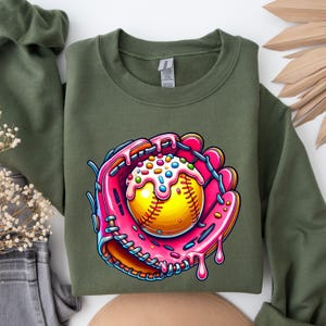 May include: A green sweatshirt with a pink and brown cartoon image of a softball in a catcher's mitt. The softball is covered in pink frosting and colorful sprinkles.