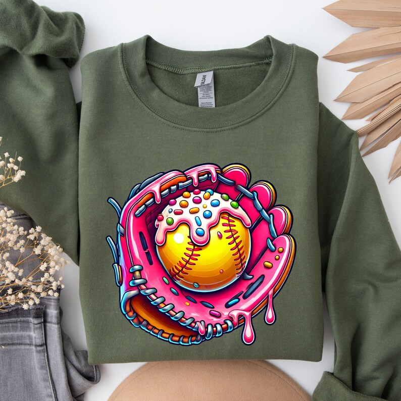May include: A green sweatshirt with a pink and brown cartoon image of a softball in a catcher's mitt. The softball is covered in pink frosting and colorful sprinkles.