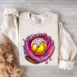 May include: A cream-colored sweatshirt with a graphic of a pink and orange baseball glove holding a yellow baseball covered in pink frosting and colorful sprinkles.