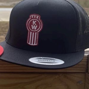 May include: Black and red trucker hat with a white and red KW logo. The hat has a black mesh back and a black snap closure. The hat is made by Yupoong.