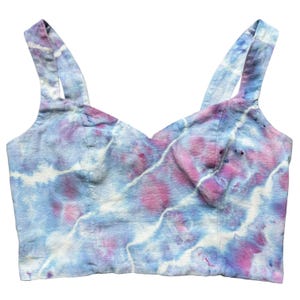 May include: A tie-dye crop top with a sweetheart neckline and thin straps. The top is made of a light blue fabric with pink and white tie-dye patterns.