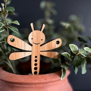 May include: A wooden dragonfly garden stake with a smiling face and a brown wood grain finish. The stake is inserted into a terracotta pot filled with green leafy plants.