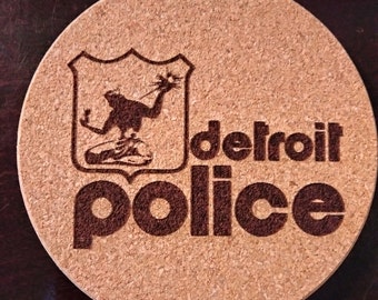 Può includere: Un sottobicchiere di sughero marrone con le parole "Detroit Police" e un'immagine stilizzata di una figura alata in uno scudo.
