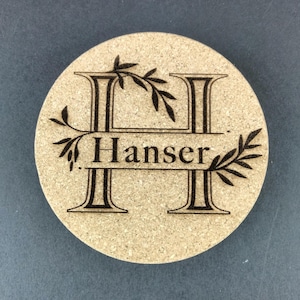 May include: Round cork coaster with a laser-etched design. The design features the letter "H" with the name "Hanser" below it, and decorative leaf accents. The coaster is light brown with a dark brown design.