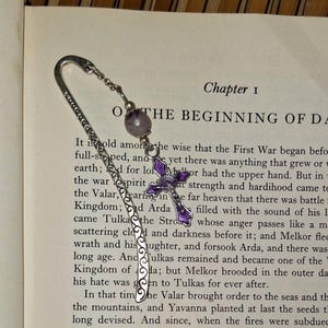 May include: A silver-toned metal bookmark featuring an intricate scrollwork pattern along its length. Attached to the top is a small silver chain holding a round, translucent purple bead and a small silver cross charm with purple enamel detailing.