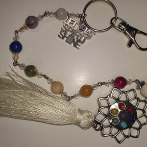 May include: A silver keychain with a white tassel and a charm featuring a silver flower with seven colorful gemstones. The charm is attached to a silver chain with clear crystals and colorful beads. The keychain also has a silver ring and a silver clasp. The charm has the text "Let Your Spirit Shine" on it.