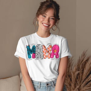 Puede incluir: Camiseta blanca con la palabra "MAMA" en letras coloridas y estampadas. Las letras están delineadas en negro y tienen un diseño juguetón y de dibujos animados. Una prenda casual para el día a día.