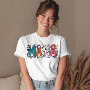 Puede incluir: Camiseta blanca con la palabra "MAMA" en letras coloridas y caprichosas. Las letras presentan varios diseños, incluyendo estampado de cebra, lunares y una flor. La camiseta es de cuello redondo.