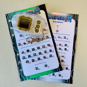 May include: A clear plastic Digimon device with a gold-colored screen and buttons, attached to a keychain. Two white cards with colorful Digimon illustrations and Japanese text are also present. The cards have a green and black border.