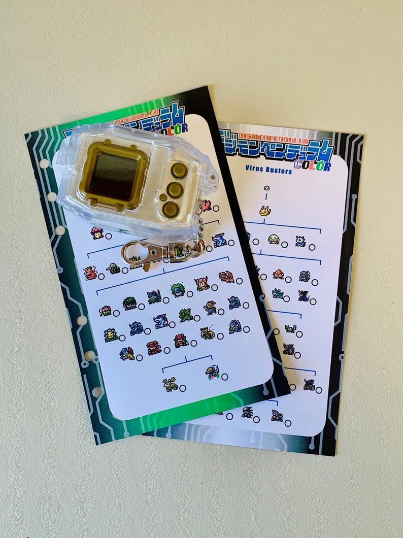 May include: A clear plastic Digimon device with a gold-colored screen and buttons, attached to a keychain. Two white cards with colorful Digimon illustrations and Japanese text are also present. The cards have a green and black border.