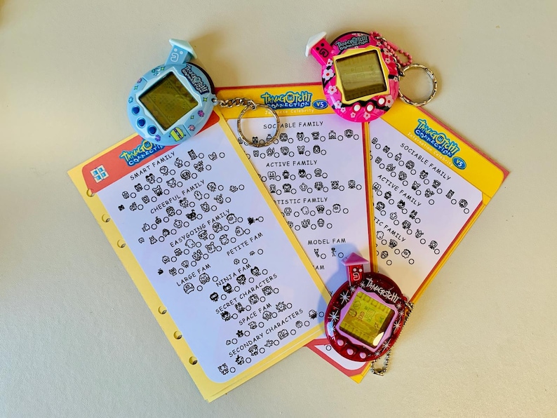 May include: A collection of vintage Tamagotchi digital pets in blue, pink, and red, with keychains, alongside yellow cards featuring character family charts. The cards include text such as "Smart Family" and "Sociable Family".
