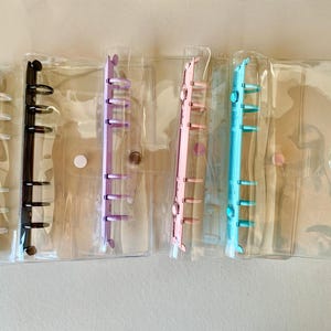 May include: Clear plastic binder organizers with colored rings in black, purple, pink, and blue. Each binder has a clear plastic cover and a snap closure. The binders are designed for organizing papers and other small items.