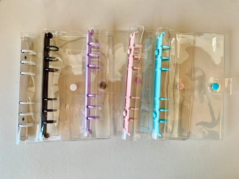 May include: Clear plastic binder organizers with colored rings in black, purple, pink, and blue. Each binder has a clear plastic cover and a snap closure. The binders are designed for organizing papers and other small items.