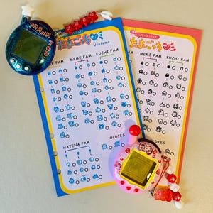 May include: Two Tamagotchi virtual pet devices, one blue and one pink, with matching charts. The blue device has the text "THANK YOU" and the pink device has a red button. The charts have Japanese text and illustrations of characters.