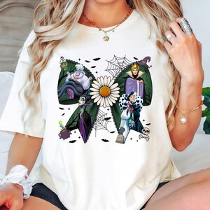 May include: White t-shirt with a green bow design featuring Disney villains, a daisy, and a spiderweb. The villains include Ursula, the Evil Queen, Maleficent, and Cruella de Vil. The design includes bats, a cauldron, and a tombstone.