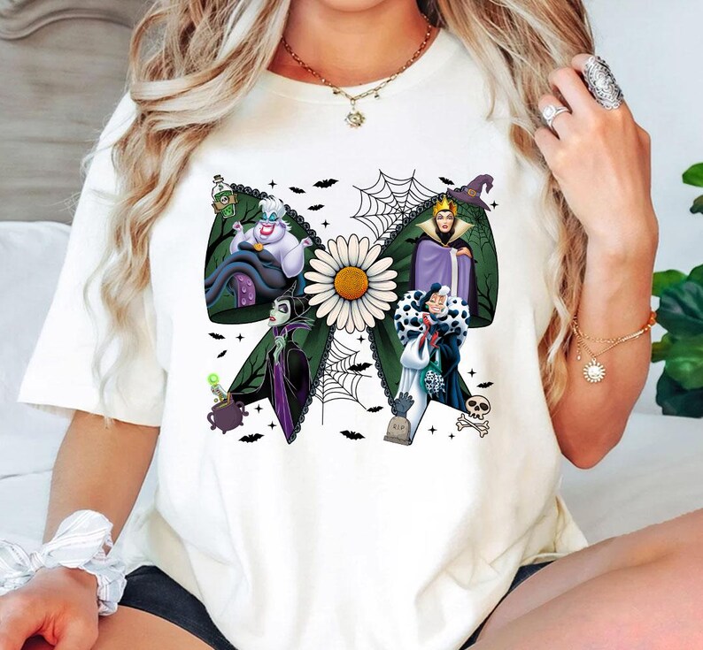 May include: White t-shirt with a green bow design featuring Disney villains, a daisy, and a spiderweb. The villains include Ursula, the Evil Queen, Maleficent, and Cruella de Vil. The design includes bats, a cauldron, and a tombstone.