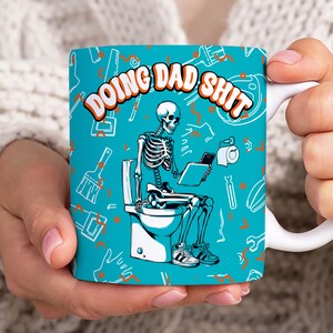 Puede incluir: Taza de cerámica blanca con fondo turquesa y la frase "DOING DAD SHIT" en naranja. La taza presenta un esqueleto sentado en un inodoro, sosteniendo una tableta y papel higiénico, con herramientas dispersas.