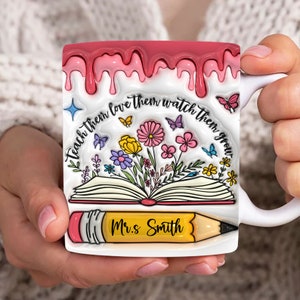 May include: White ceramic coffee mug with a pink drip design. The mug features an illustration of an open book with flowers and butterflies. The text "Teach them love them watch them grow" is written around the book. A yellow pencil with the text "Mrs. Smith" is on top of the book.