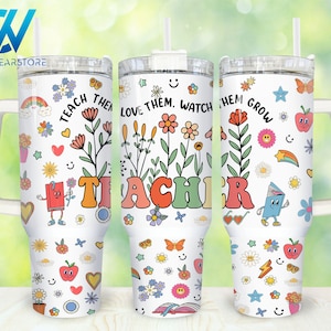 Puede incluir: Tres vasos de acero inoxidable blancos con tapas y asas. Cada vaso presenta un diseño diferente con flores coloridas, corazones, estrellas, arcoíris y las palabras "Teach Them Love Them Watch Them Grow" y "Teacher".