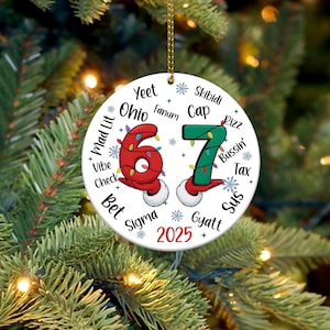 May include: A white ceramic Christmas ornament with the numbers "67" in red and green, decorated with Christmas lights. The ornament has various slang terms and the year "2025" printed around the numbers. It hangs from a gold string.
