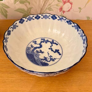 Può includere: Ciotola in ceramica bianca con bordo smerlato e disegni blu di fiori e rami di alberi. La ciotola presenta un'illustrazione circolare centrale di un albero e uccelli. Il bordo è decorato con motivi floreali blu.