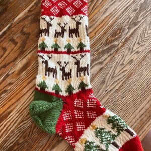 Peut inclure: Une chaussette de Noël en tricot rouge et verte avec un fond blanc. La chaussette présente un motif de rennes, de sapins et de diamants.