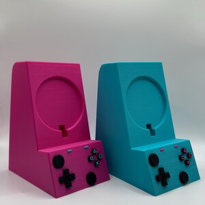 May include: Two 3D-printed game controller stands, one magenta and one teal. Each stand features a circular holder, a slot, and a controller layout with buttons and a directional pad. The stands are designed to hold a game controller.