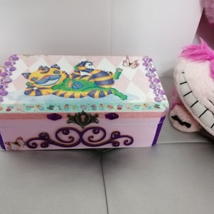 Peut inclure: Une boîte en bois rose avec une bordure violette et une illustration fantaisiste du Chat du Cheshire d'Alice au Pays des Merveilles. La boîte a un loquet en métal décoratif et est ornée de paillettes et de strass.