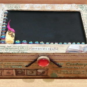 Könnte beinhalten: Eine Holzkiste mit einem Scharnierdeckel und einem kleinen, roten, runden Knopf. Die Kiste hat ein schwarzes Schwarzes Brett im Inneren mit einem kleinen goldenen Schlüssel und einer rosa Feder. Die Kiste ist mit verschiedenen Bildern und Texten verziert, darunter "Adventures Await" und "New Country".