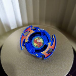 May include: A blue and orange Beyblade with a red and yellow phoenix design on the center piece. The Beyblade is called "Drazi-S".