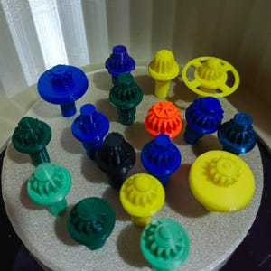 以下が含まれることがあります： 様々な形とサイズのカラフルな3Dプリントされたこまのコレクション。青、緑、黄色、オレンジ、黒の色合いです。歯車のようなデザインのものもあれば、平らまたは丸い表面のものもあります。
