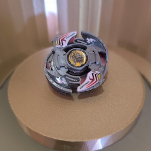 May include: A silver and red Beyblade with a yellow and black centre. The Beyblade has the word "DRAGON" written on it.