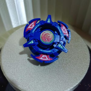 May include: Blue and red plastic Beyblade with a red and white dragon design on the center. The Beyblade has the text "Dranzer GT" and "Gronzer GT" on it.
