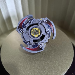 May include: A grey and red Beyblade with a yellow and black design. The Beyblade has the text "DRIGER G" on the side.