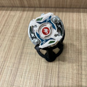 May include: A white and blue Beyblade with a red and white center. The Beyblade is on a black stand. The Beyblade has a silver rim and green accents.