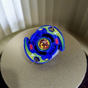 May include: A blue and green Beyblade with a red and yellow dragon design in the center. The Beyblade is on a light brown surface.