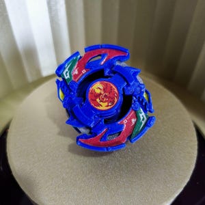 May include: A blue and red Beyblade with a dragon design. The Beyblade has a green and white sticker on the side that says "Dragoon G".