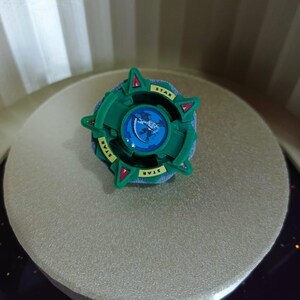 Op de afbeelding: Een groene en blauwe plastic speelgoedtol met een stervormig ontwerp en het woord "STAR" erop gedrukt. De tol heeft een blauw centrum met een wit en zwart schepselontwerp.