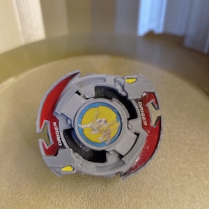 Peut inclure: Un Beyblade gris et rouge avec un motif jaune et bleu au centre. Le Beyblade porte l'inscription "DRIGER".