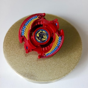 Op de afbeelding: Een rood en geel speelgoed met de tekst "DEATH GARGOYLE" is gecentreerd op een goudkleurig cirkelvormig oppervlak. Het speelgoed heeft een centraal ontwerp met een zwart figuur. Het speelgoed heeft een drietandig ontwerp met blauwe accenten.