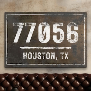May include: A rectangular wall art piece with the text "77056 HOUSTON, TX" in white, distressed font. The background is a dark, textured brown, framed with a white border. The artwork is displayed above a brown leather sofa.