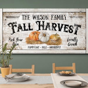 May include: A rectangular wooden sign with a white frame. The sign reads "The Wilson Family Fall Harvest" with illustrations of pumpkins. Text includes "Pick Your Own" and "Locally Grown". The sign is on a table.