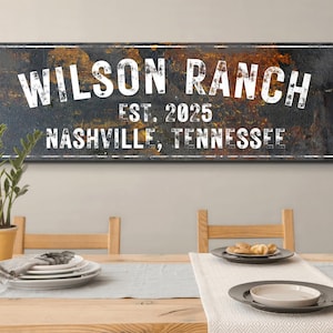 May include: A rectangular wooden sign with the words "WILSON RANCH EST. 2025 NASHVILLE, TENNESSEE" in white lettering. The sign has a distressed, textured background with dark and gold tones. The sign is displayed above a dining table.
